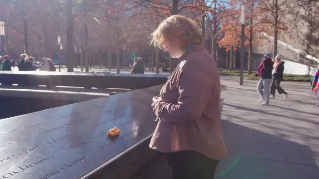 Un fotograma del documental muestra a una mujer rubia con un taxi amarillo de juguete frente al memorial de las víctimas del 11-S