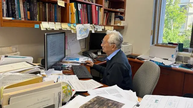 Hiroo Kanamori trabajando en su oficina en Caltech en setiembre de 2026. Está sentado frente al computador, su mesa está cubierta por documentos y sismogramas