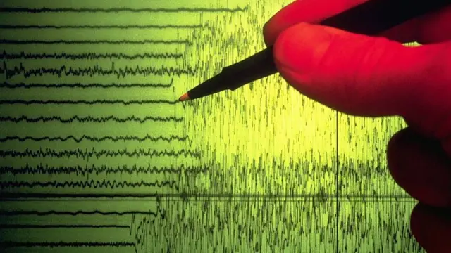 Imagen de las ondas registradas en un sismograma. Una mano con un lápiz apunta al punto donde se ve un cambio en la traza de las ondas debido al inicio de un terremoto.